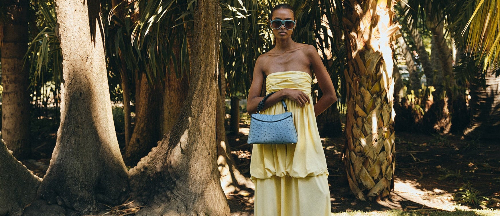 Woman in a yellow dress holding a blue rope handle ostrich leather handbag in a tropical setting with palm trees.