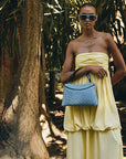 Woman in a yellow dress holding a blue rope handle ostrich leather handbag in a tropical setting with palm trees.